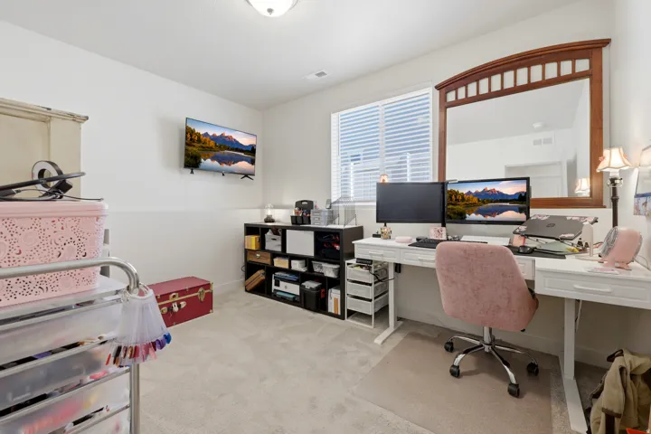 Bedroom (currently used as an office space) featuring a large window with horizontal blinds, a mounted television, a built-in desk, and light-colored carpeting