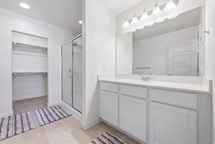 Spacious bathroom featuring a double vanity with white cabinetry and light-toned countertops, a glass-enclosed shower, and a dedicated walk-in closet with shelving and hanging rods