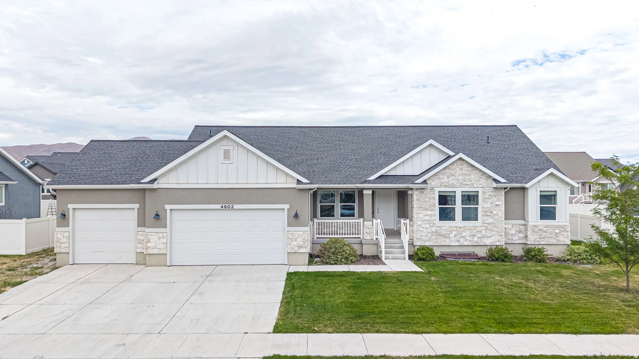 Ranch-style residence featuring a gray exterior with stone accents, a dark shingle roof, a two-car garage with an additional single-car garage, and a covered front porch with white railing