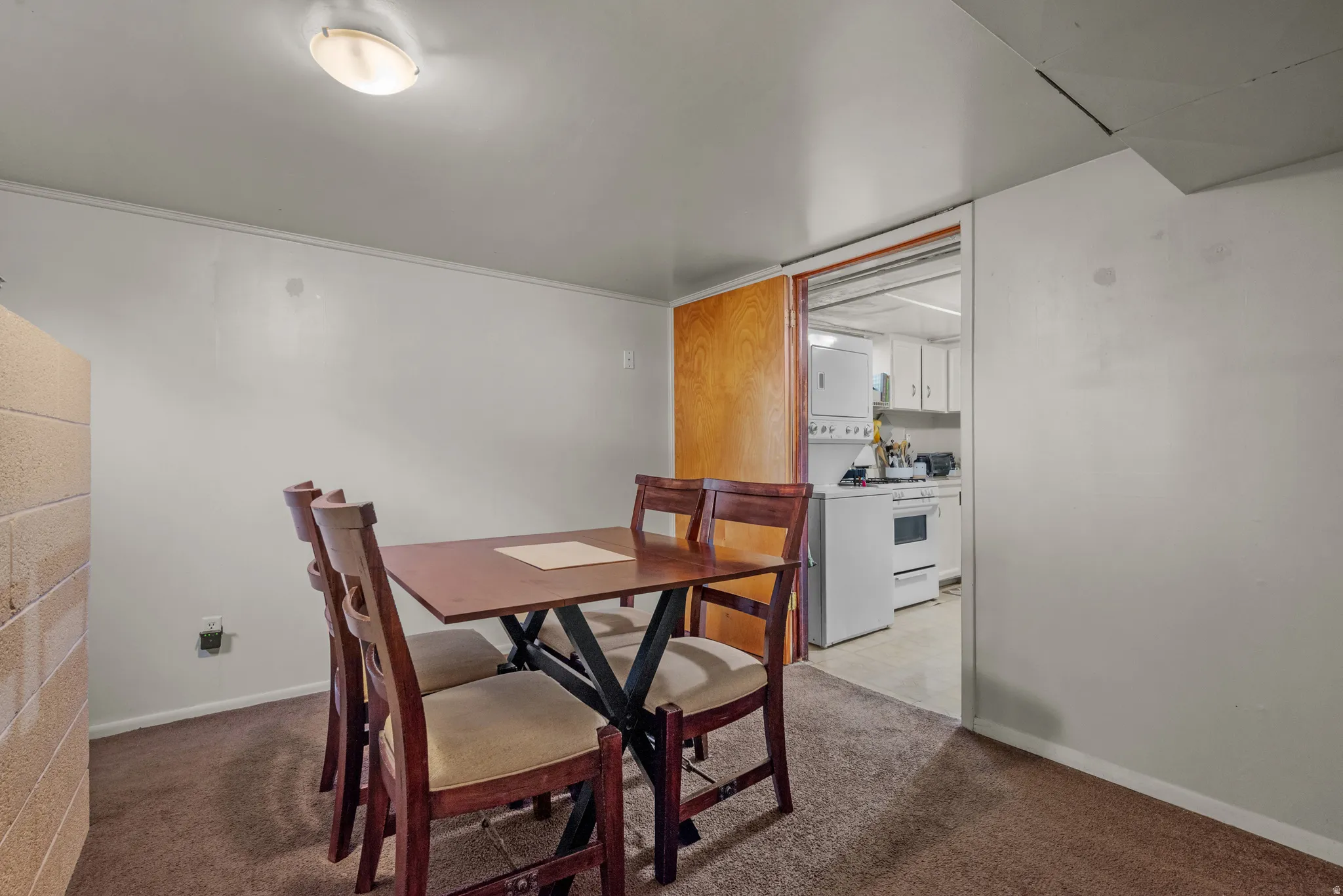 Carpeted dining area featuring white walls, a single ceiling light fixture, and a sliding wood-finish door revealing a partial kitchen with white cabinetry and appliances