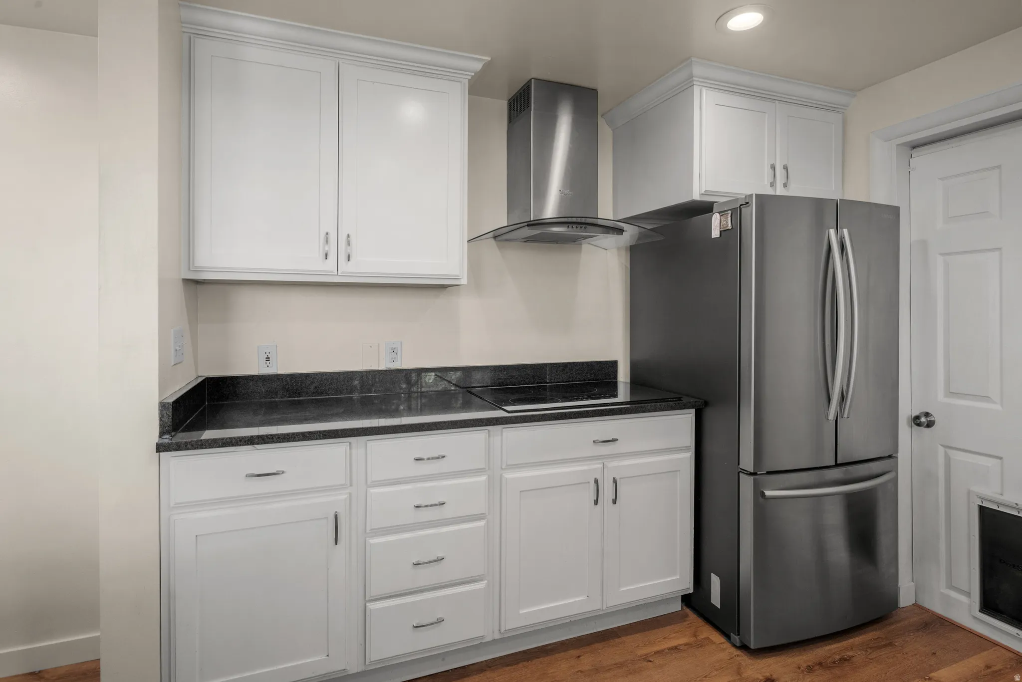 Kitchen featuring white cabinetry with brushed nickel hardware, black countertops, a stainless steel range hood, and a stainless steel refrigerator