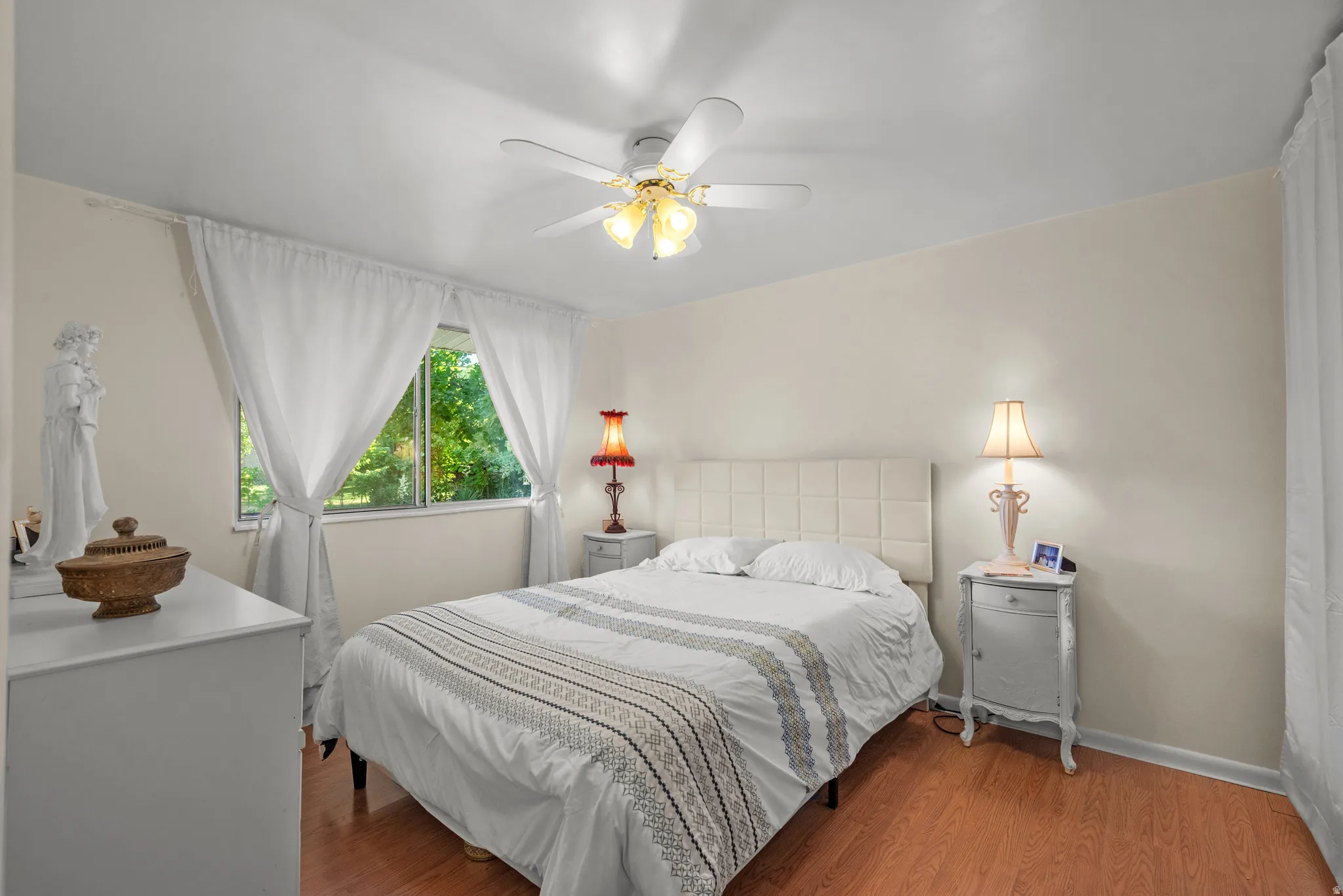 Bedroom featuring wood-finish flooring, a ceiling fan with integrated lighting, and a window with white drapes