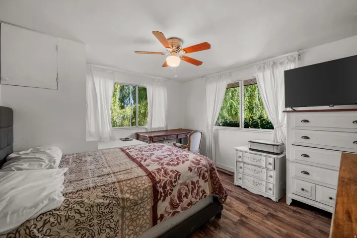 Bedroom featuring wood-finish flooring, two window openings with white trim, and a ceiling fan with integrated lighting