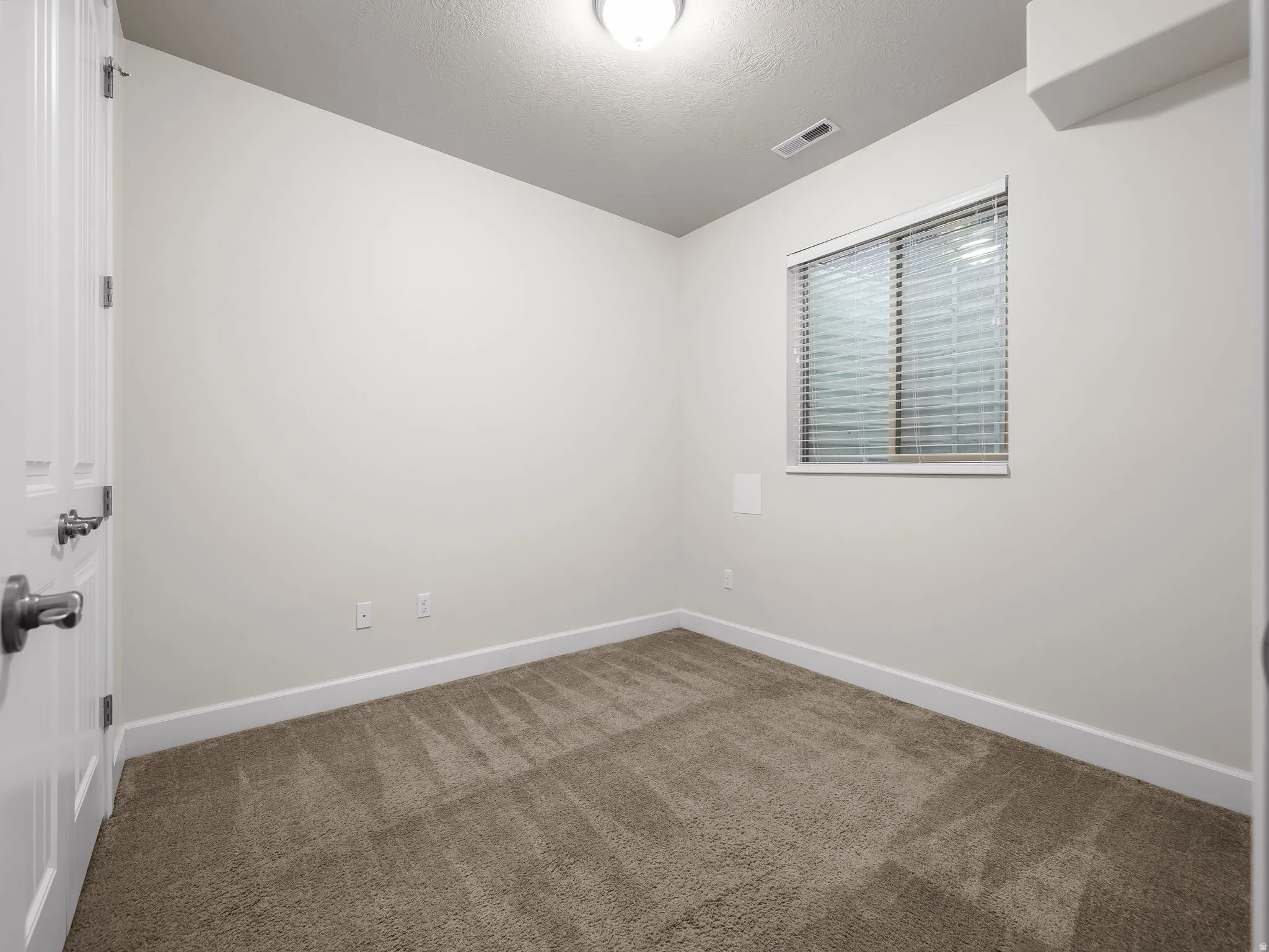 Neutral-toned room featuring wall-to-wall carpeting, a single window with horizontal blinds, and a flush-mount ceiling light fixture