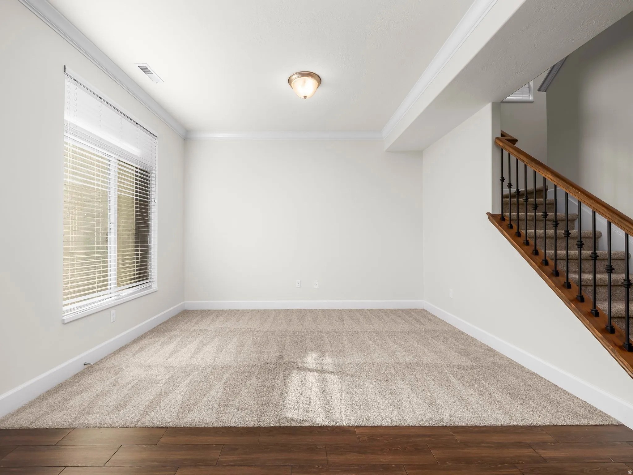 Versatile room featuring wood-finish flooring transitioning to patterned carpet, a large window with blinds, and a staircase with wood handrails and wrought iron balusters