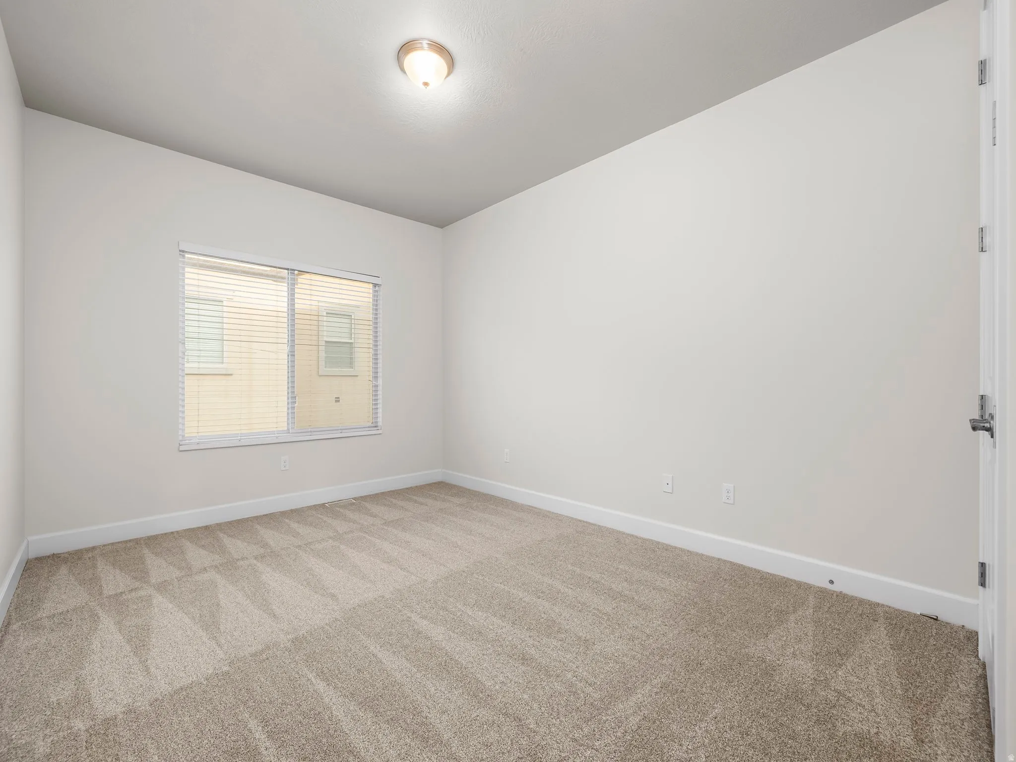 Neutral-toned room featuring a window with horizontal blinds, overhead lighting, and patterned carpet flooring