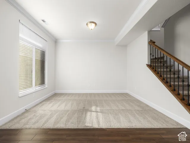 Versatile room featuring wood-finish flooring transitioning to patterned carpet, a large window with blinds, and a staircase with wood handrails and wrought iron balusters