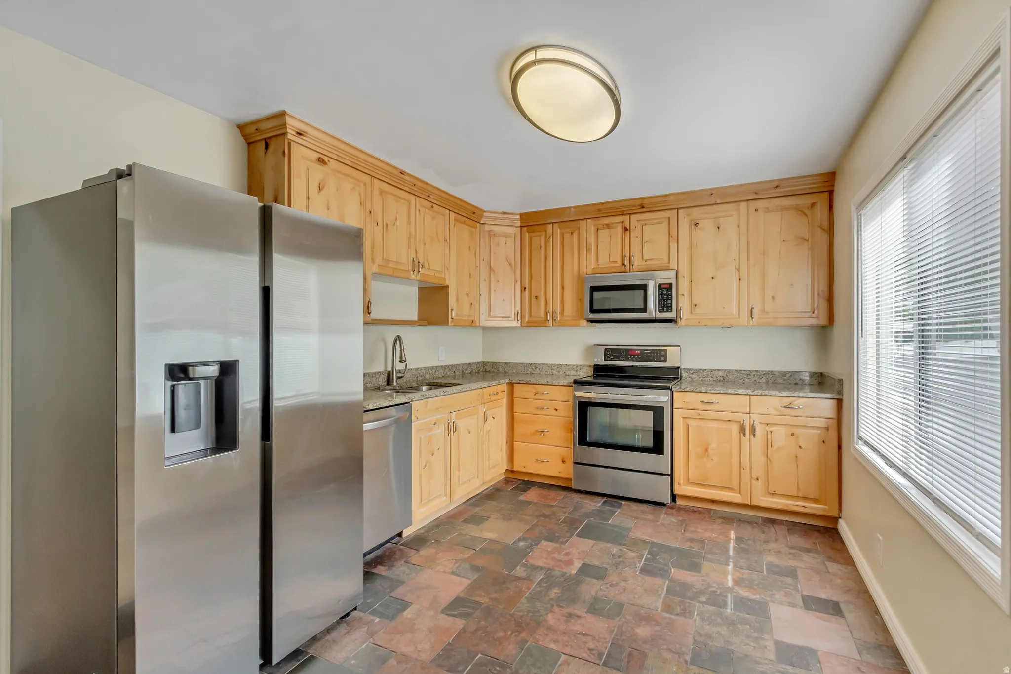 Kitchen featuring light wood cabinetry, stainless steel appliances, speckled countertops, and multi-tone tile flooring