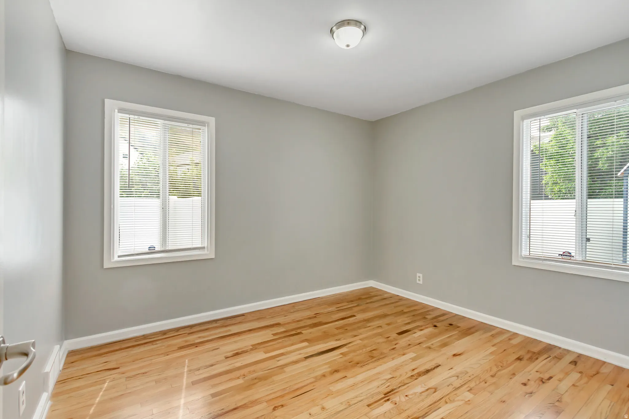 Bright room featuring wood-finish flooring, two windows with blinds, light gray wall paint, and white trim