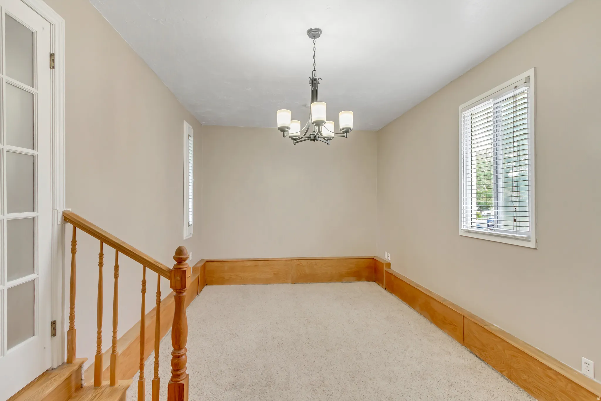 Carpeted room featuring a ceiling-mounted chandelier, natural wood half-wall paneling, and two windows with blinds