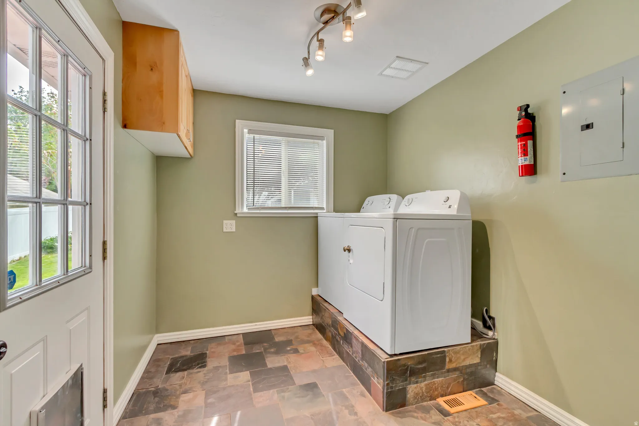 Utility area featuring slate tile flooring, a window with blinds, a wood-finish wall cabinet, and track lighting