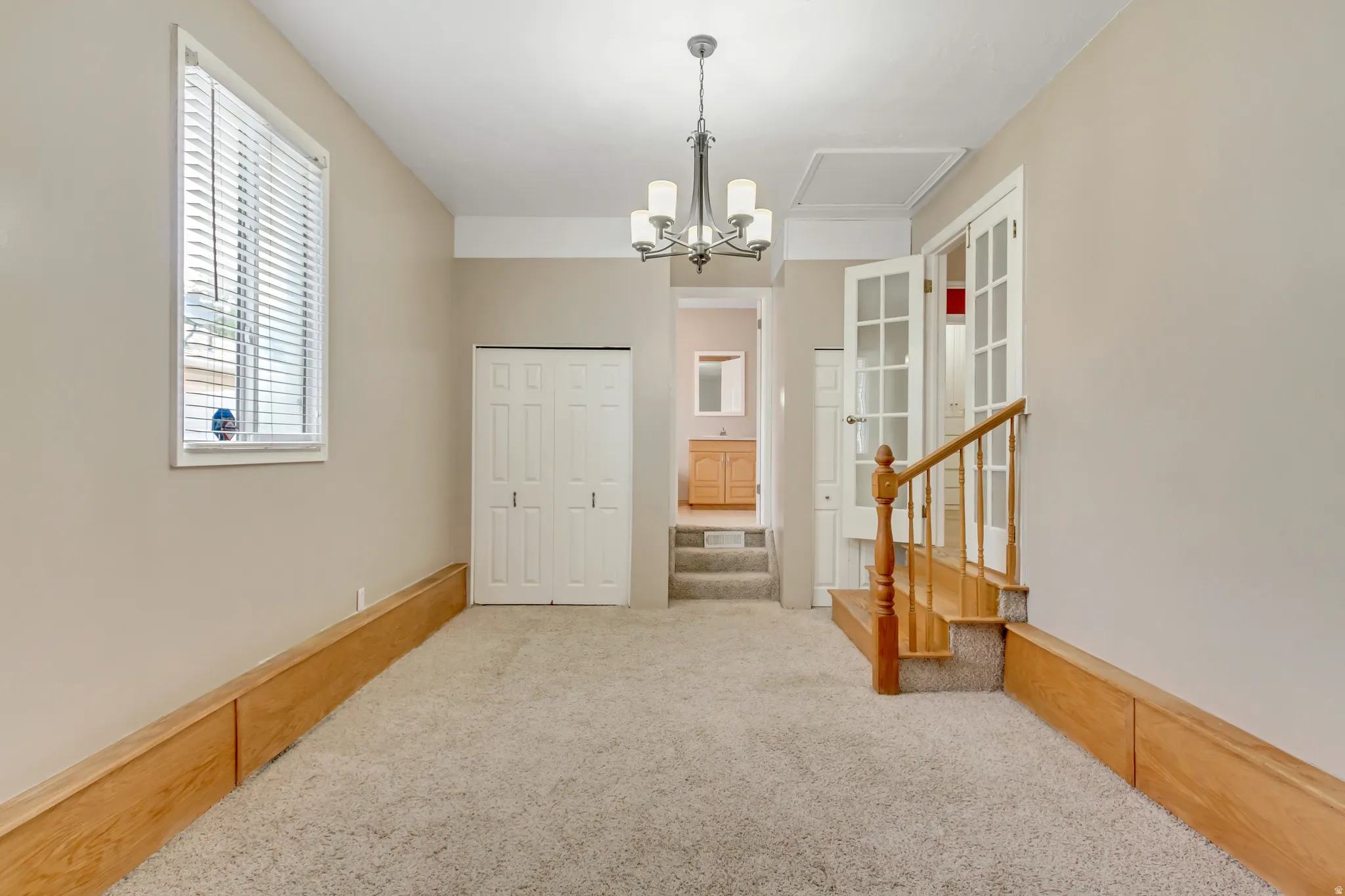 Carpeted room with neutral wall tones, featuring a single window with blinds, bi-fold closet doors, a chandelier, and a wood-finish staircase with a railing