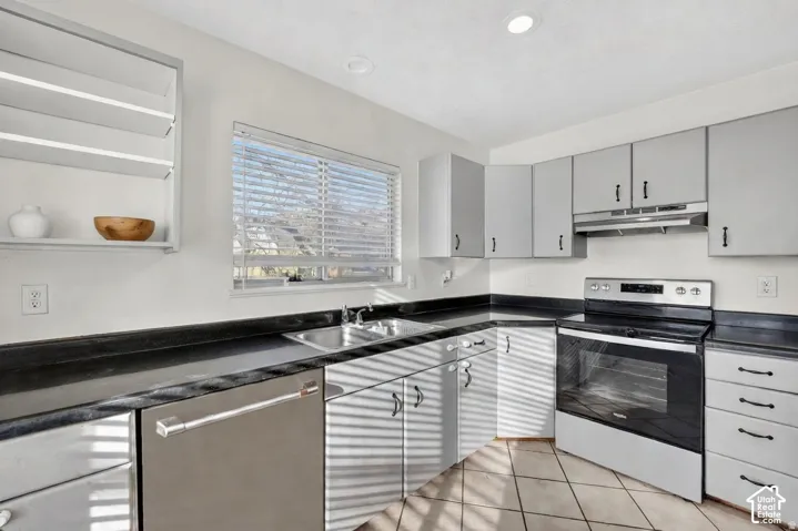 Corner kitchen featuring gray cabinetry, a stainless steel dishwasher, black countertops, a stainless steel sink, and a white oven with a black cooktop
