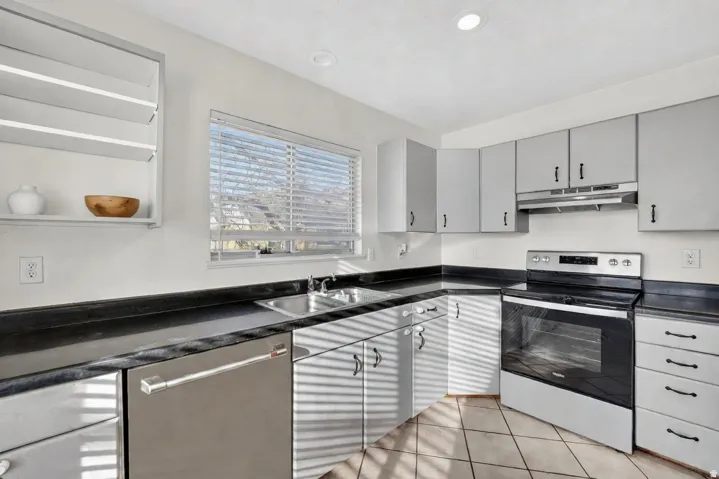 Corner kitchen featuring gray cabinetry, a stainless steel dishwasher, black countertops, a stainless steel sink, and a white oven with a black cooktop