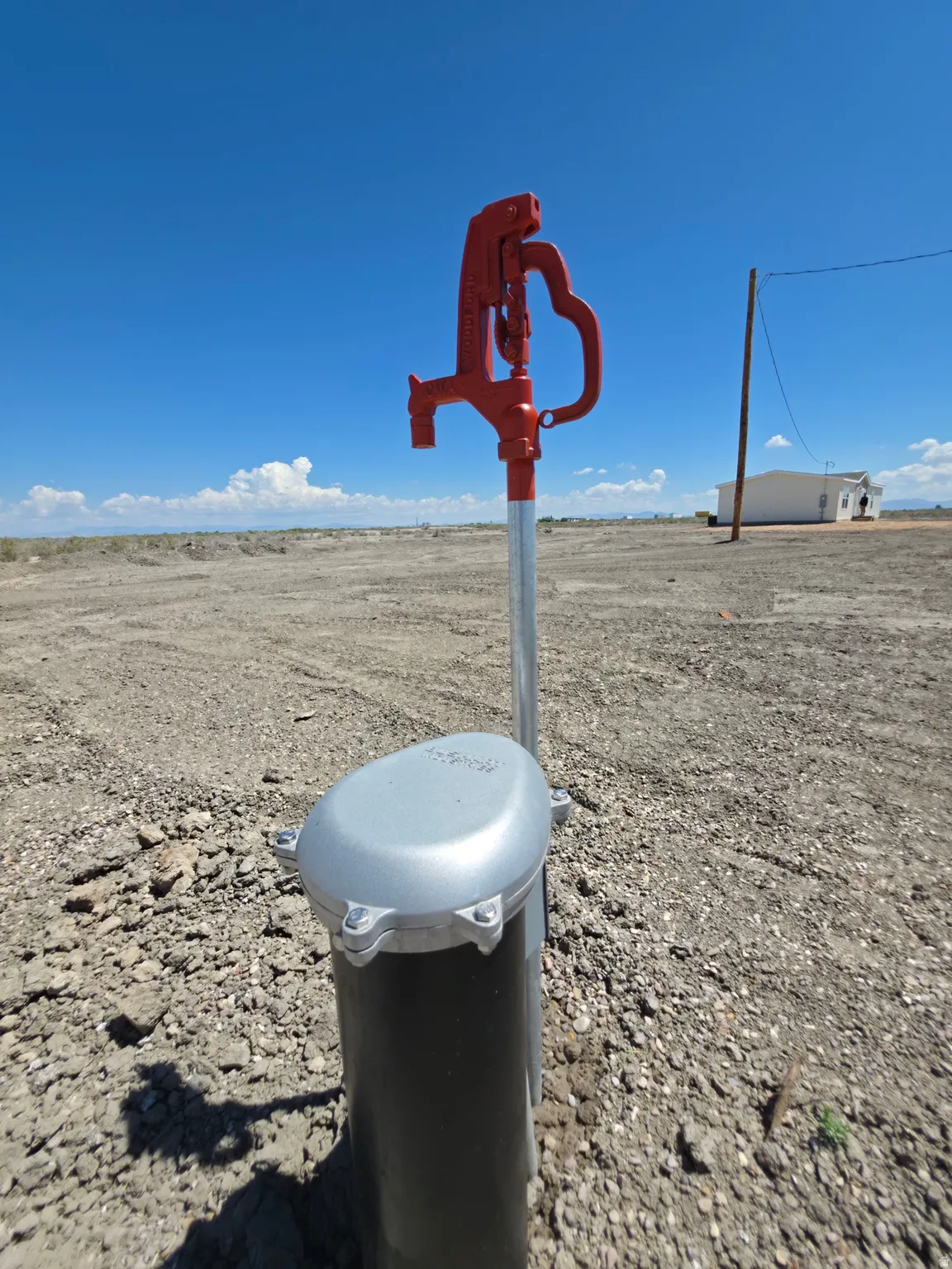 Red manual pump with a galvanized steel standpipe and a gray well cap, set against a clear blue sky