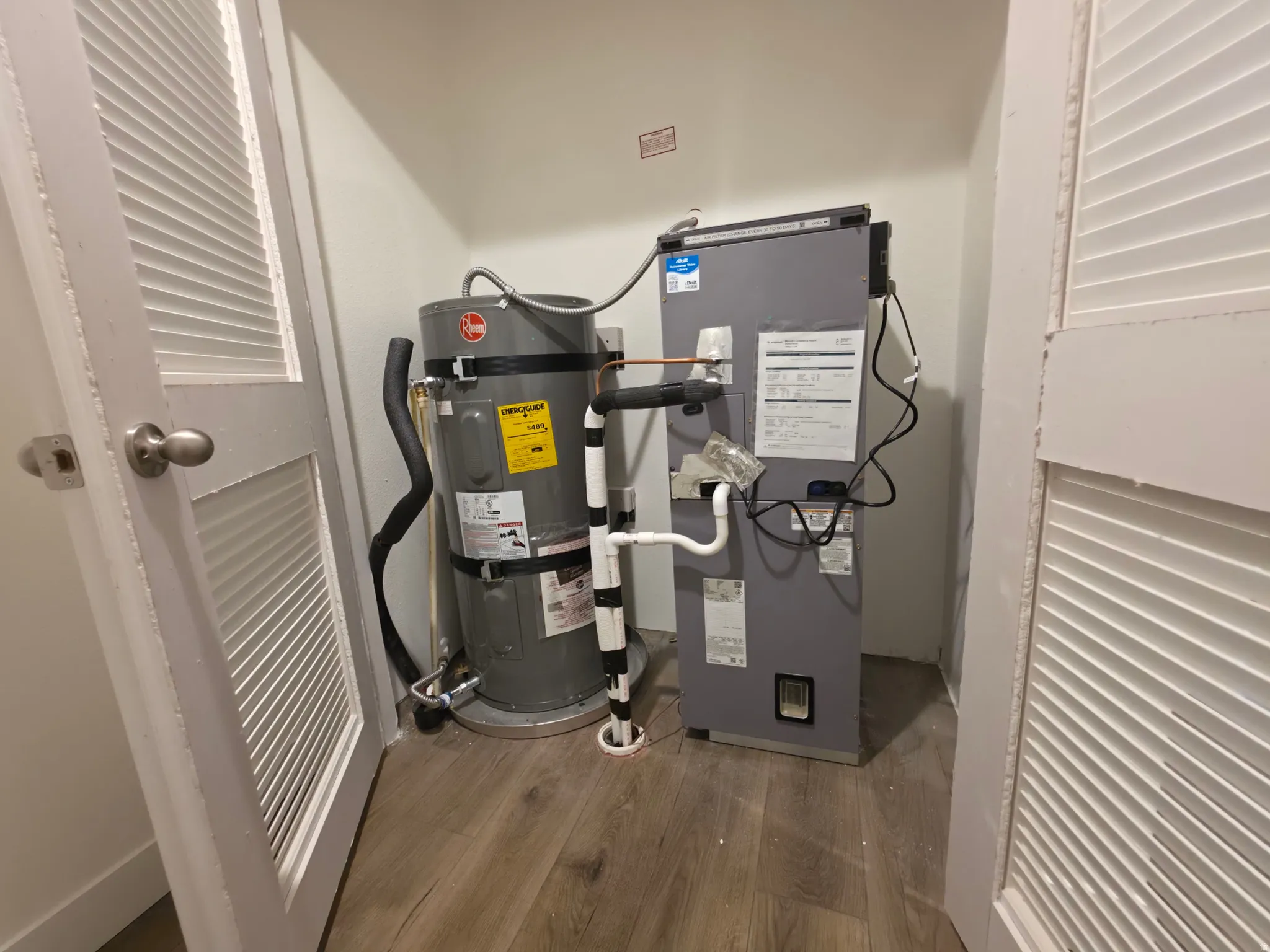 Utility closet featuring water heater and air handler unit, hard surface flooring, and louvered bifold doors