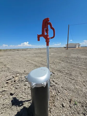 Red manual pump with a galvanized steel standpipe and a gray well cap, set against a clear blue sky