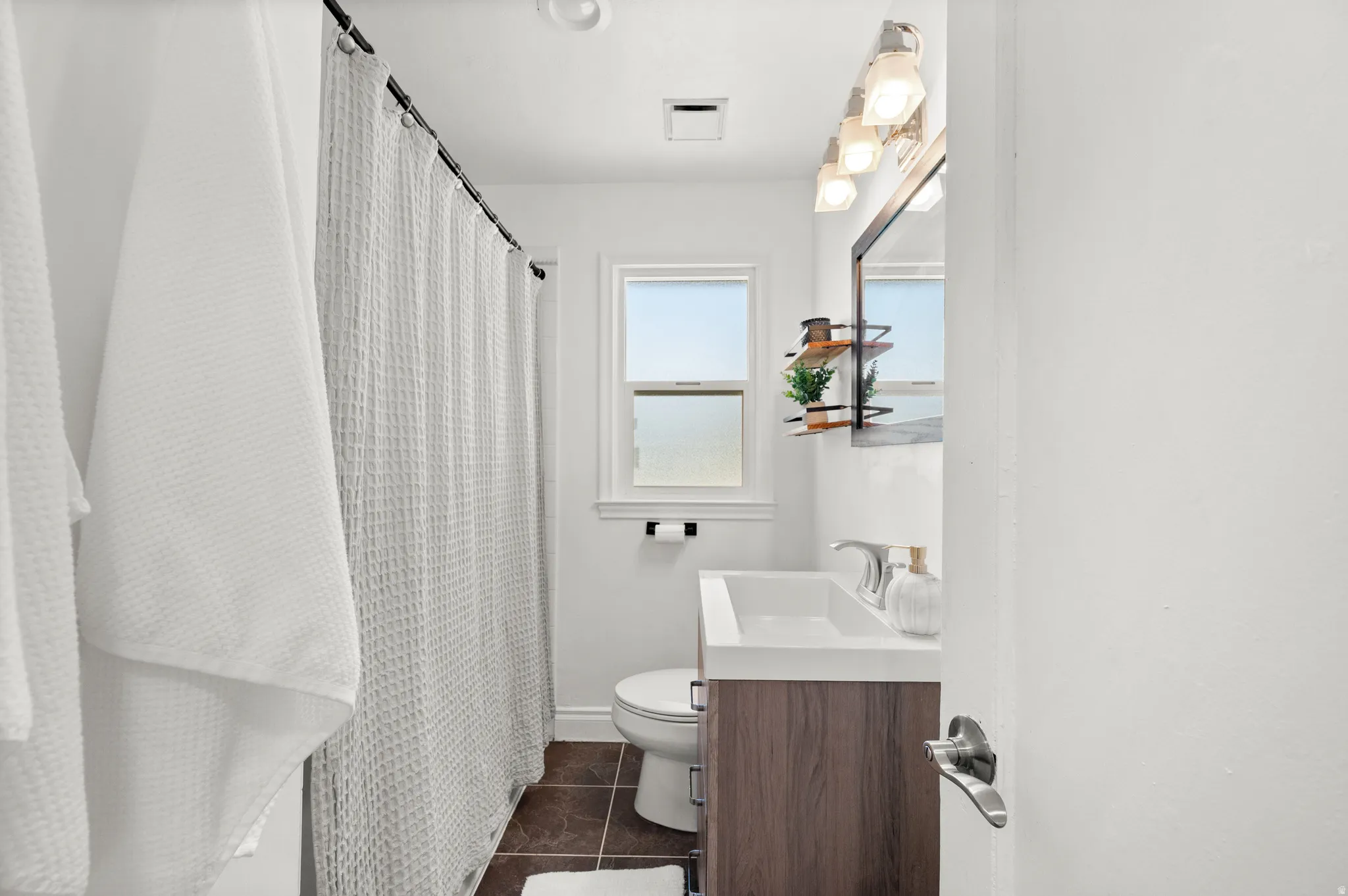 Full bathroom featuring a single window, a dark wood-finish vanity with an integrated sink, a wall-mounted mirror, and tile flooring