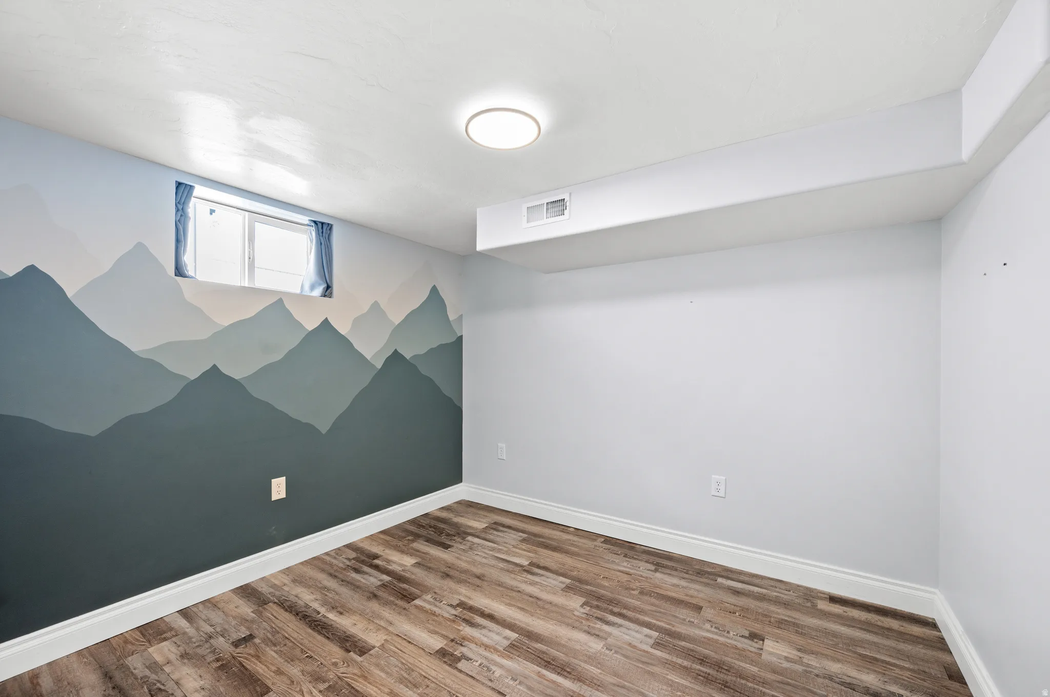 Room featuring wood-finish flooring, a mountain-themed accent wall, a flush-mount ceiling light, a window with blue curtains, and white baseboards