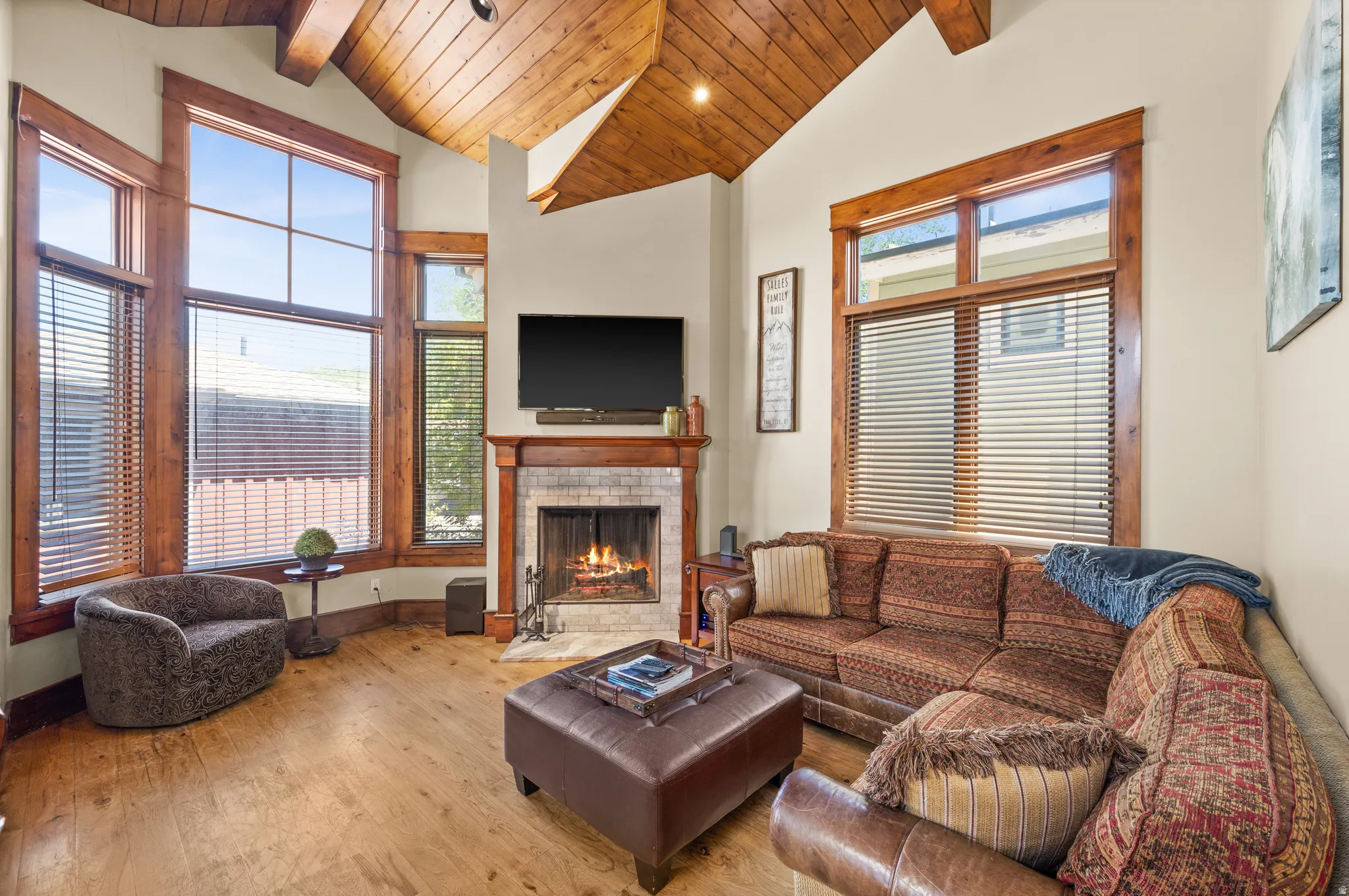Living area featuring vaulted wood-finish ceilings with exposed beams, wood-finish flooring, a stone-surround fireplace with a wood mantel, and expansive multi-pane windows with wood-finish trim