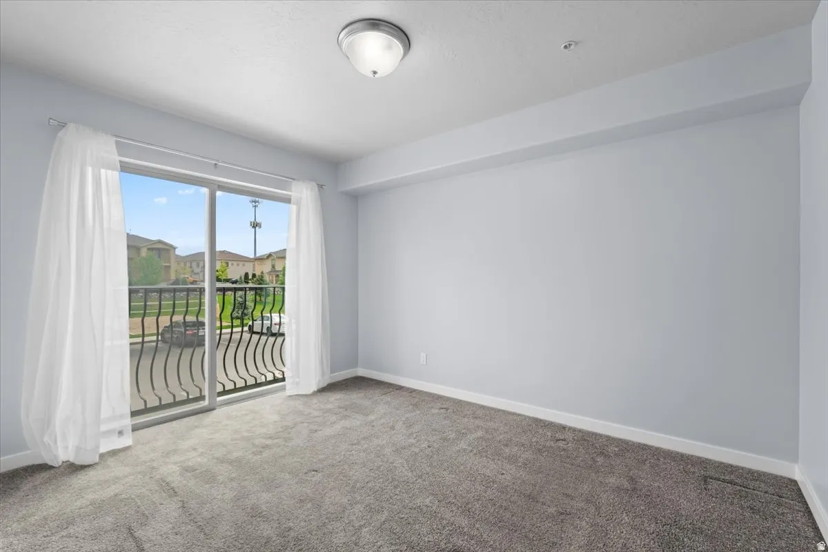 Spacious room with sliding glass doors opening to a balcony with black wrought iron railing, featuring light gray walls, gray carpet flooring, and a flush mount ceiling light
