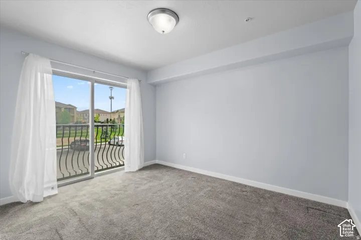 Spacious room with sliding glass doors opening to a balcony with black wrought iron railing, featuring light gray walls, gray carpet flooring, and a flush mount ceiling light