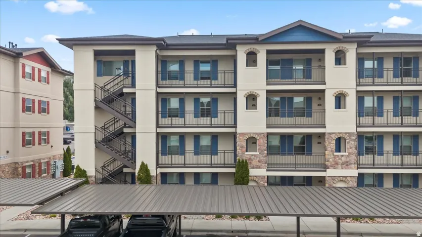 Multi-level building with stone and stucco exterior, featuring individual balconies with black railings, blue window shutters, and a covered parking structure