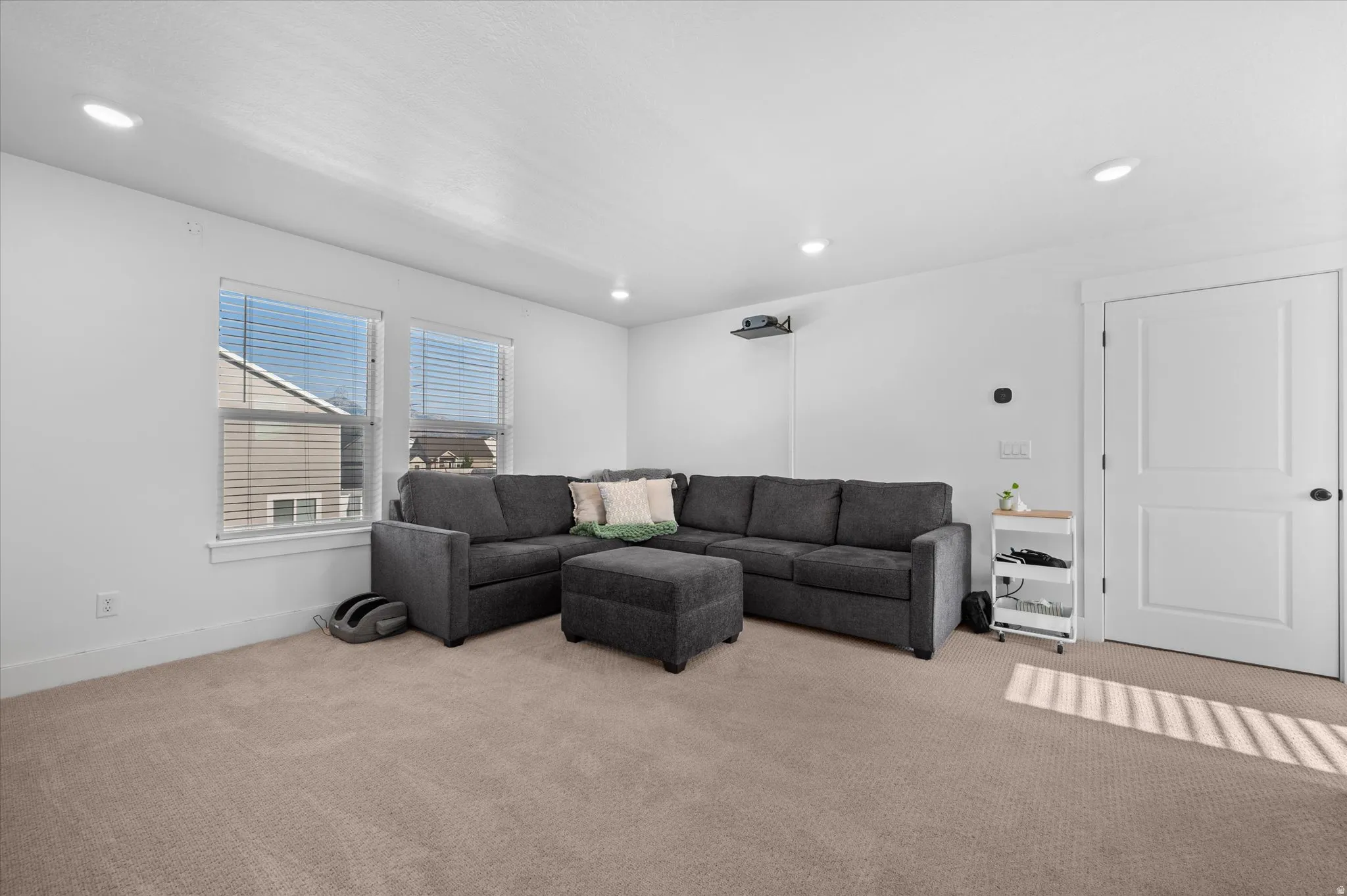 Recessed ceiling lighting, pair of windows with blinds, panel interior door with dark hardware, and wall-mounted projector