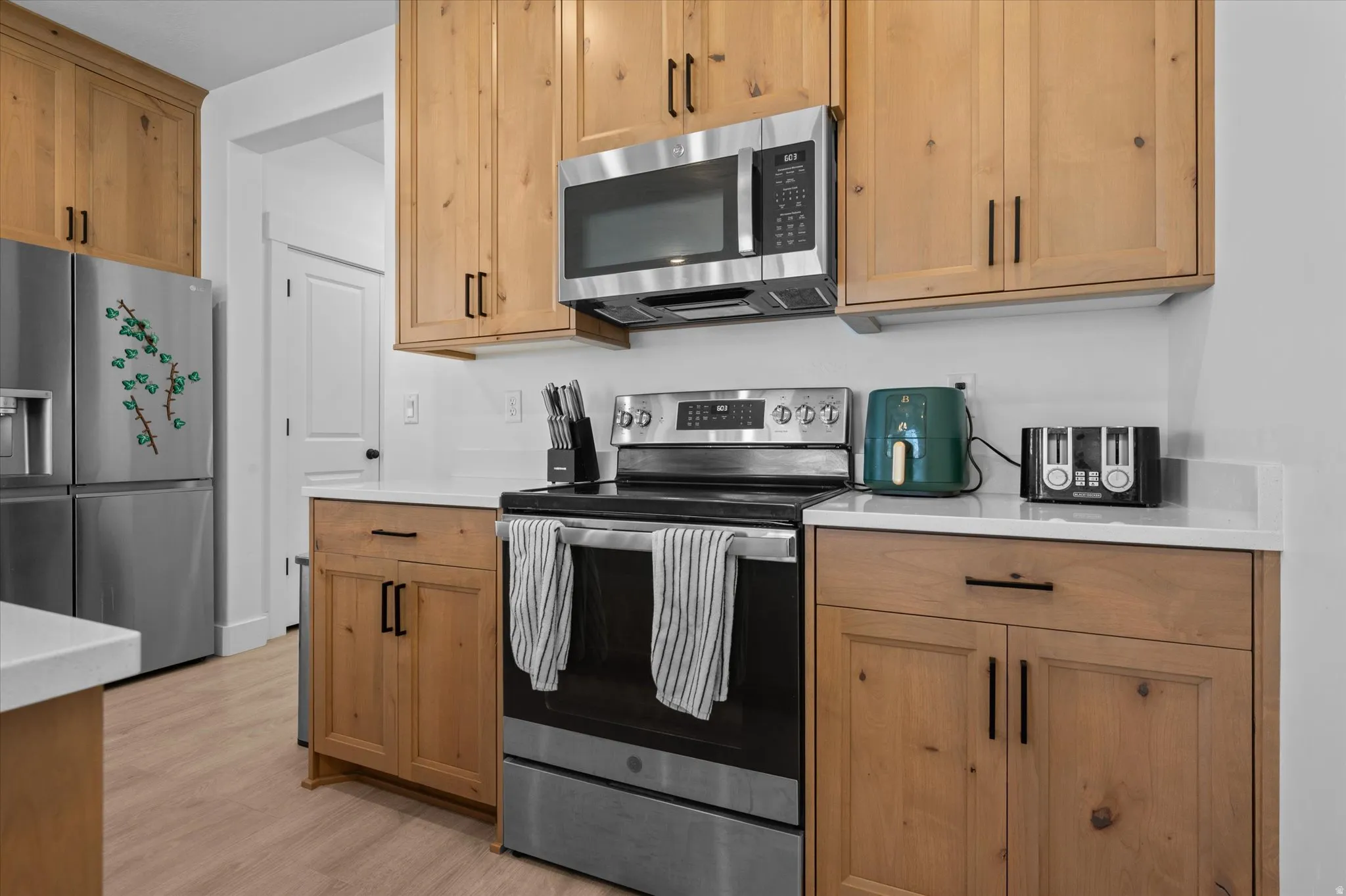 Light wood-finish cabinetry with black hardware, stainless steel appliances, white countertops, and wood-finish flooring