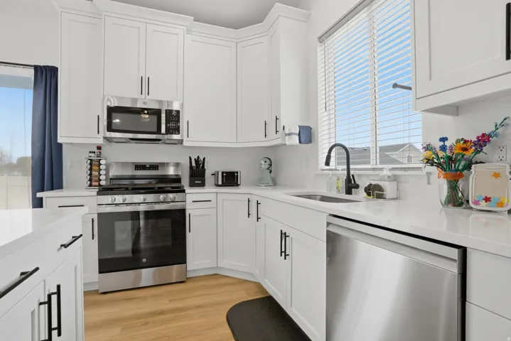 Modern kitchen featuring white shaker cabinetry with matte black hardware, stainless steel appliances, a stainless steel sink with a matte black faucet, wood-finish flooring, and white countertops