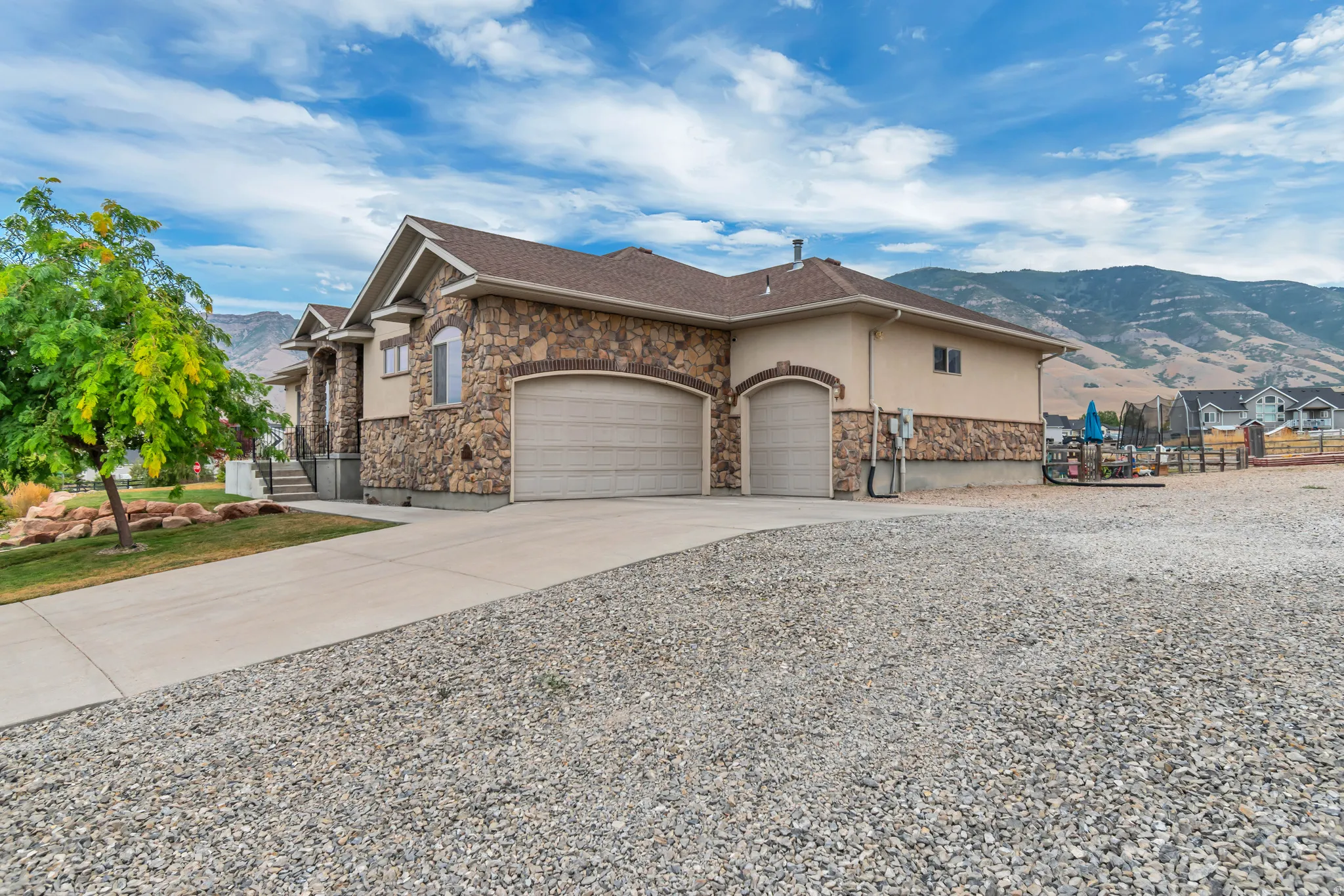 Stone and stucco exterior with a multi-car garage, concrete driveway, and gravel parking area