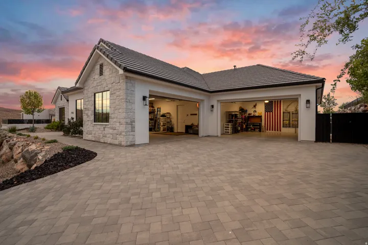 Paved driveway with a three-car garage, white stucco exterior, stone accent facade, black frame windows, and a dark tile roof