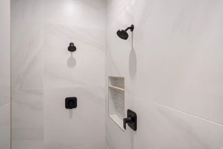 Shower enclosure with large format white tile, matte black shower heads, matte black temperature controls, and a tiled shower niche with a decorative mosaic back