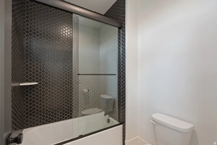 Bathtub with a black arabesque tile surround, clear glass sliding shower doors, a built-in shower shelf, and a white toilet