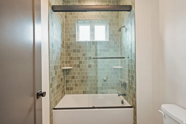 Tub and shower combination featuring a glass enclosure, square green wall tiles, and a window