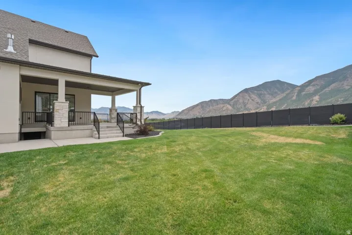Expansive rear yard featuring a manicured lawn, covered patio with stone-wrapped columns, black metal railings, and a solid perimeter fence