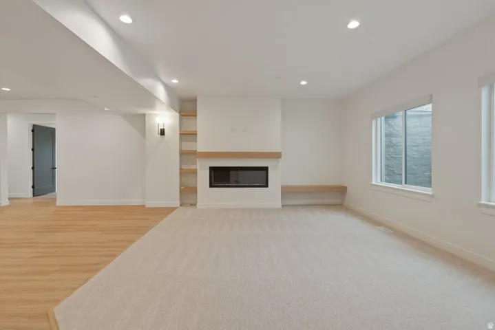 Open living area featuring a modern fireplace with a light wood mantel, recessed lighting, built-in shelving, light wood-finish flooring, and light-toned carpeting