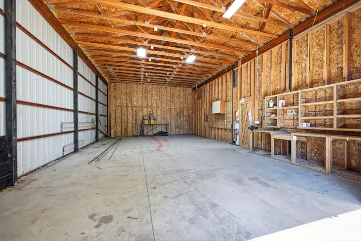 Spacious workshop area with concrete flooring, exposed wood beam ceiling, built-in shelving, and workbenches