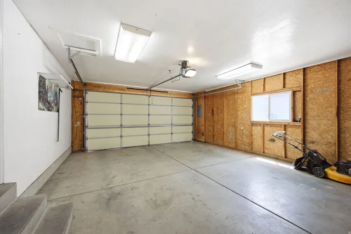 Spacious garage featuring concrete flooring, a sectional garage door, multiple fluorescent light fixtures, a window with horizontal blinds, and exposed oriented strand board walls