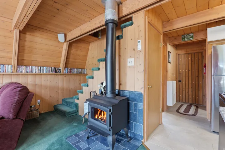 Interior space featuring a black wood-burning stove with a blue tile surround and hearth, wood paneled walls, exposed wood beams, and a partial staircase with green carpeted treads