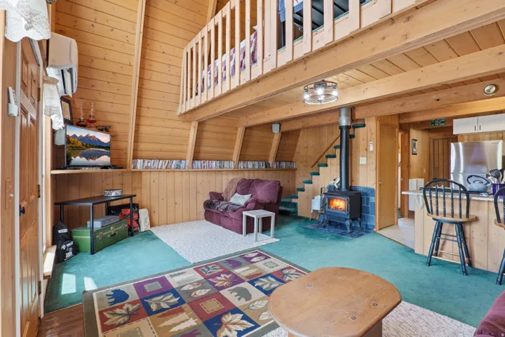 Open-concept living area featuring extensive wood paneling, a wood-burning stove with a blue tile surround, and a loft overhead with a wood railing