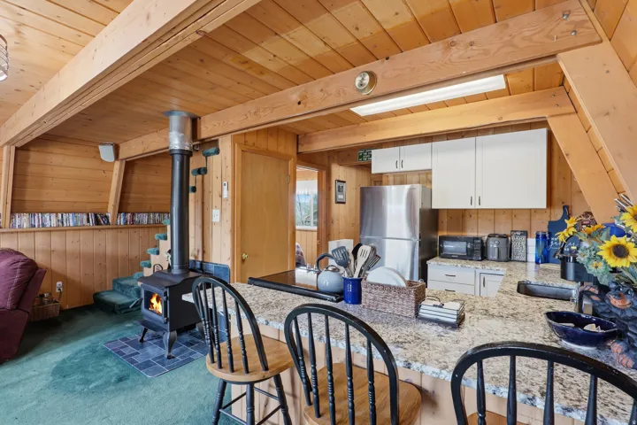 Open-concept space featuring extensive wood paneling, exposed wood beam ceiling, and a prominent wood-burning stove with a tiled hearth