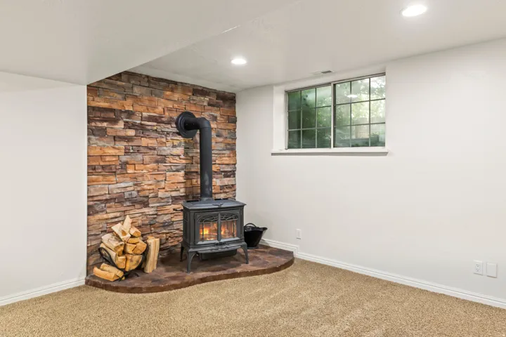 Stone veneer accent wall featuring a black wood-burning stove with a hearth, recessed ceiling lighting, a single window, and carpet flooring