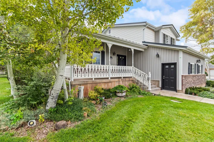 Exterior featuring light gray vertical and horizontal siding, a covered porch with white railing, black entry doors, and mature landscaping with established trees