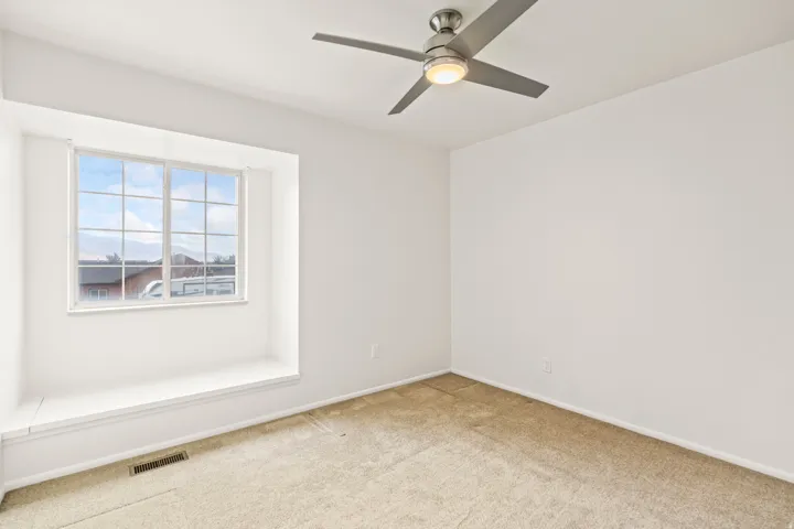 Light-filled room featuring a broad window with a built-in window seat, a modern ceiling fan with integrated lighting, clean white walls, and neutral-toned carpeting