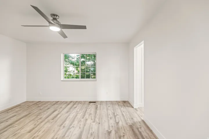 Bright room featuring wood-finish flooring, a contemporary ceiling fan with integrated lighting, white walls, and a window with exterior greenery