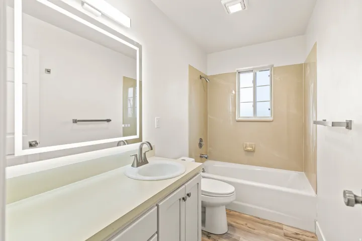 Bathroom featuring a light wood-finish floor, built-in bathtub with light-toned wall surround, and a vanity with a white countertop and integrated sink