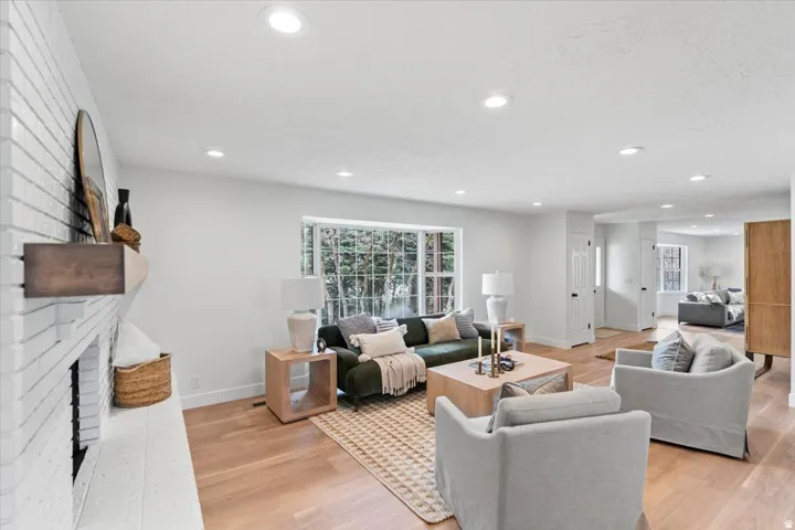 Spacious living area featuring light wood-finish flooring, a painted brick fireplace with a wood mantel, recessed lighting, and multiple windows providing natural light