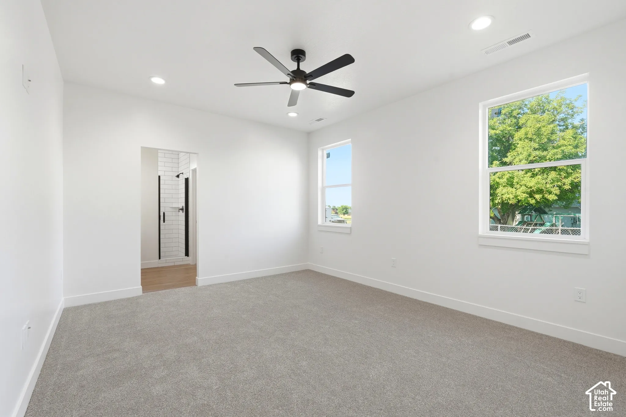 Recessed lighting, ceiling fan, two large windows, light gray carpet, and wood-finish flooring visible in the entry