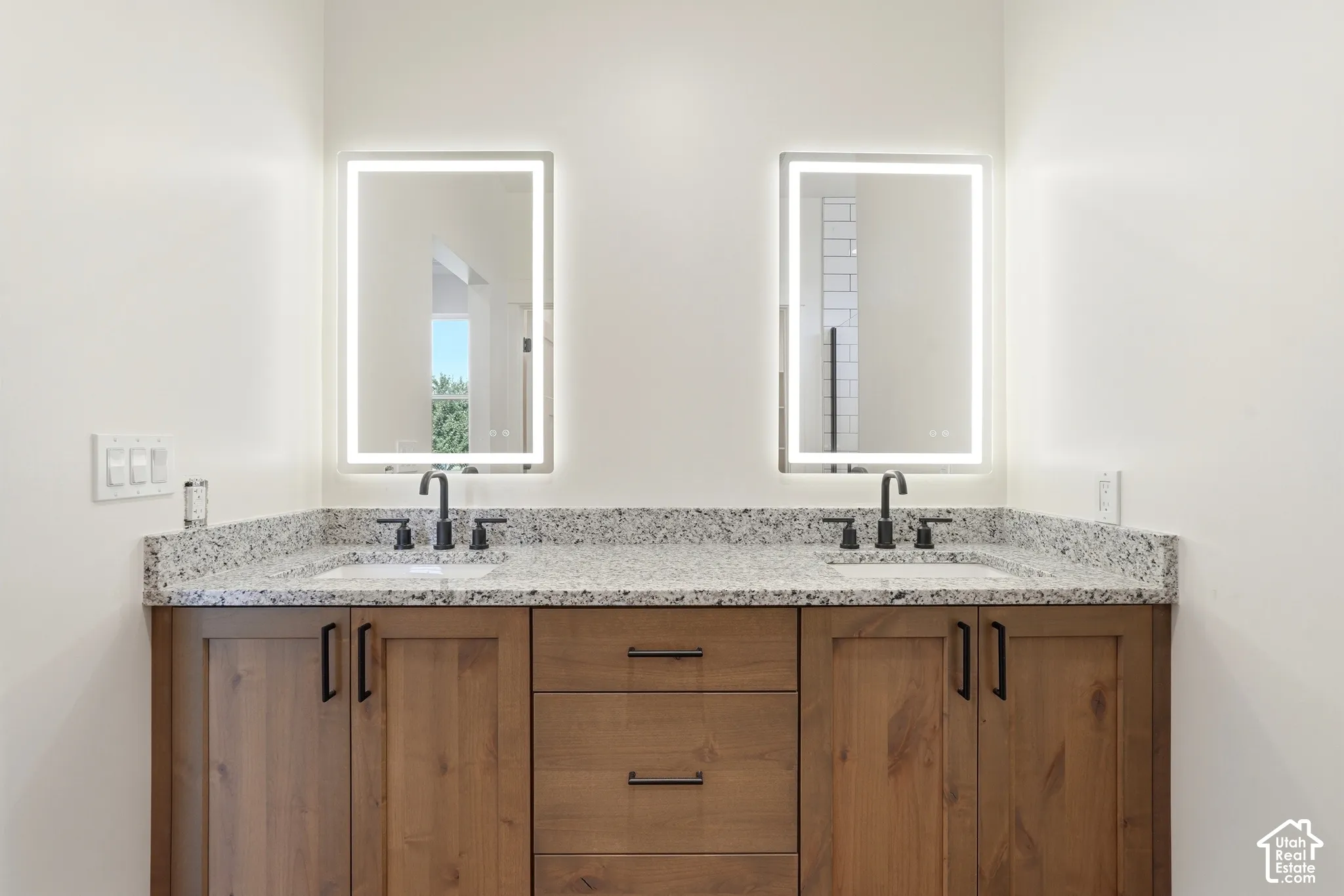 Dual vanities featuring individual lighted mirrors, wood-finish cabinetry, black hardware, and speckled countertops